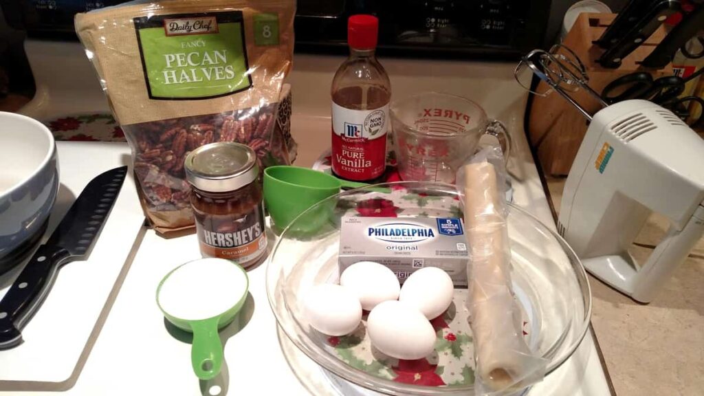 ingredients for pie: nuts, vanilla, sugar, eggs, cream cheese, jar of caramel ice cream sauce, pie crust