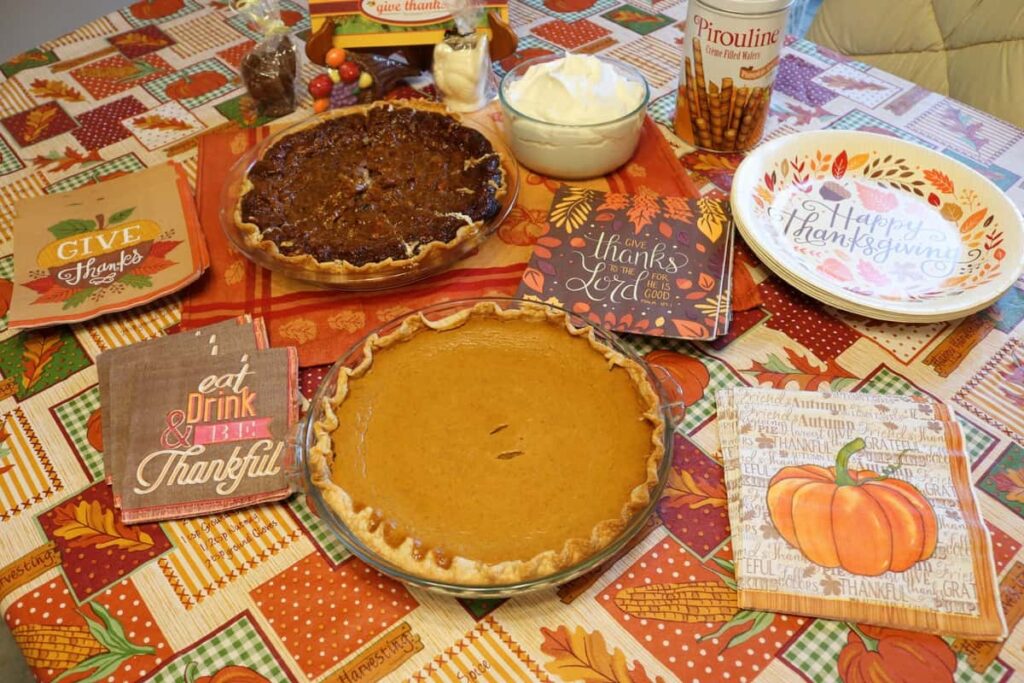 whole pumpkin pie, whole pecan pie, variety of Thanksgiving themed napkins and paper plates with bowl of whipped cream and tin of wafer rolls