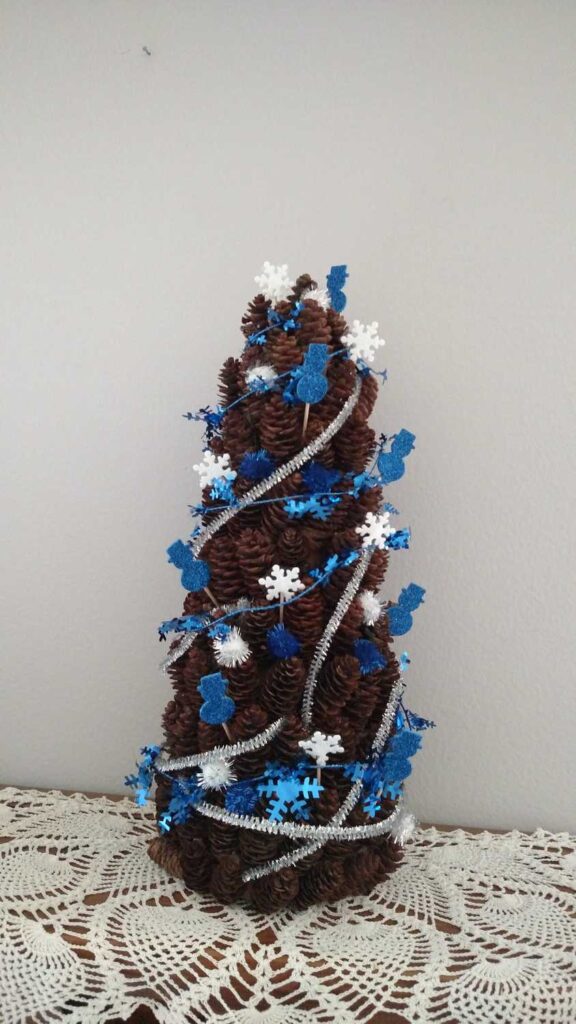 pine cone tree decorated with blue glitter snowmen, silver metallic pipe cleaners, white snowflakes, blue metallic garland, and blue pom poms