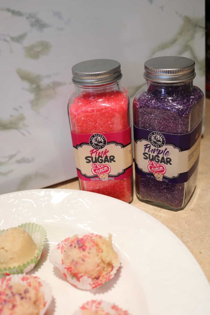 the pink and sugar sprinkles glass jars in the background with edible sugar cookie dough in foreground