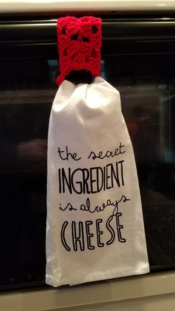 crochet towel holder holding a white towel that says "the secret ingredient is always cheese"