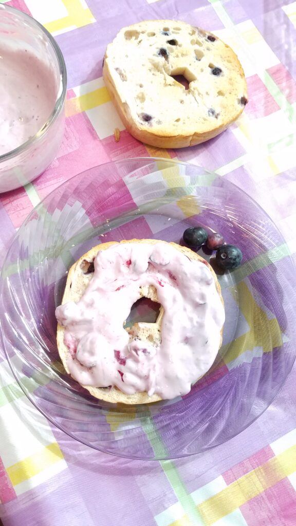 blueberry cream cheese on a bagel on a glass plate, plain blueberry bagel slice in background