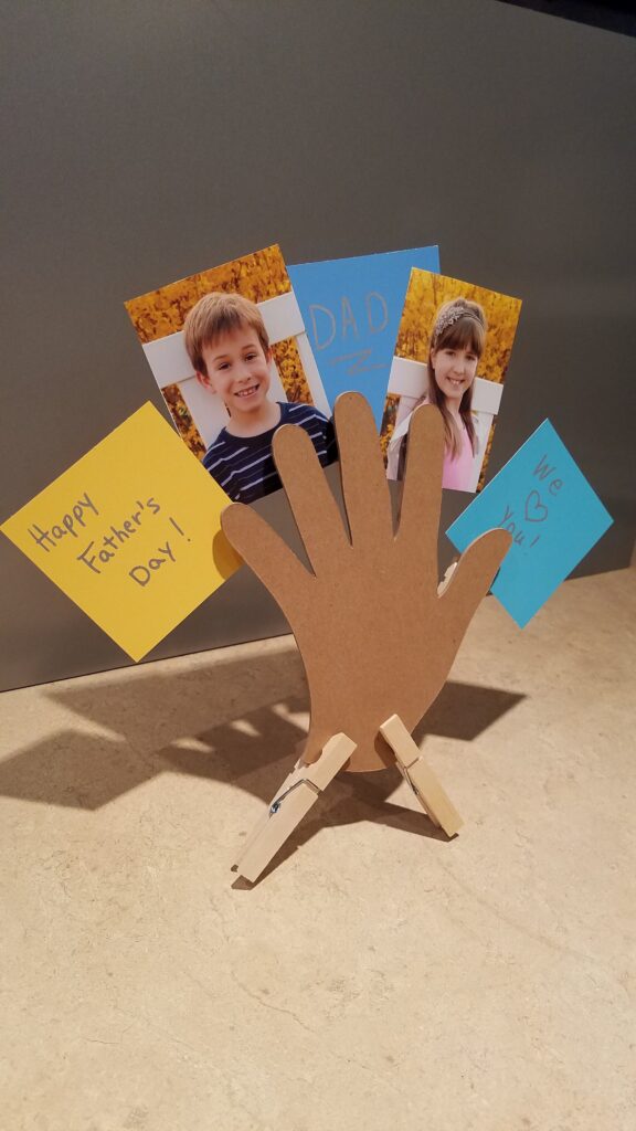 vertical picture of cut out hand standing on countertop holding children's pictures and notes to dad