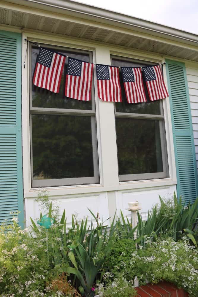 five flags hanging in front of windows on house with flowerbed underneath