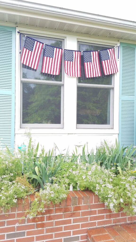 five flags hanging in front of windows on house with flowerbed underneath